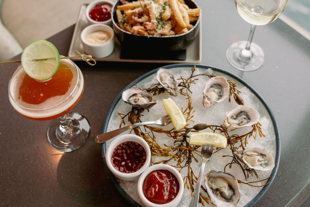 A plate of oysters on ice with condiments and a cocktail and wine glass on a table