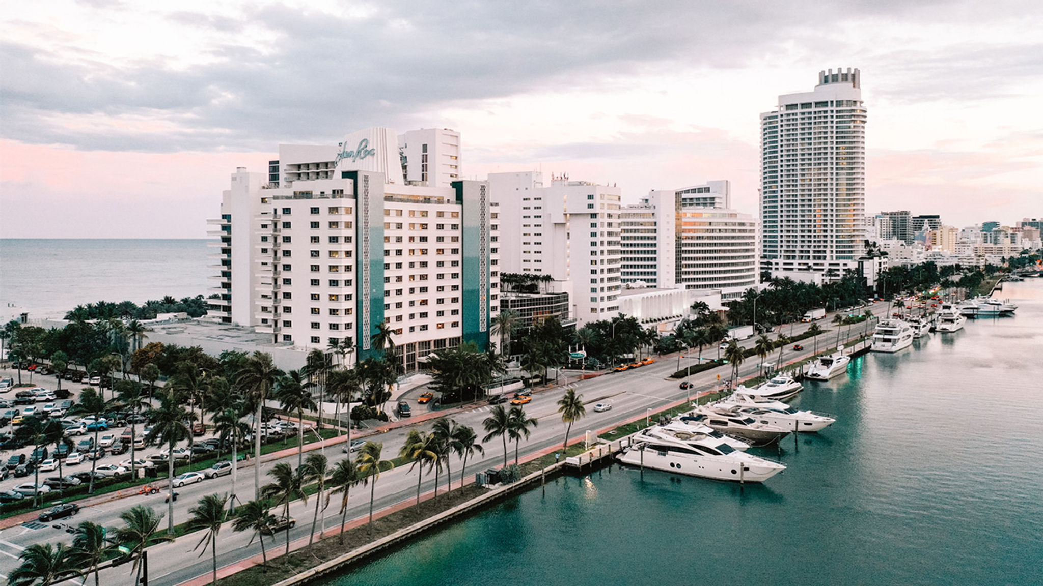 A wide exterior view of a hotel and surrounding buildings along a waterfront with boats.
