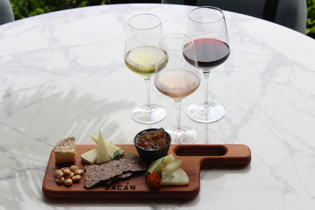 A wine and cheese board on a marble table with three wine glasses and a wooden cheese board with various cheeses, breads, and nuts.