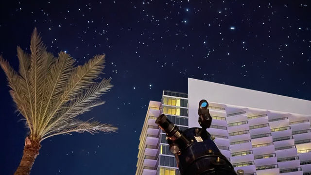 A nighttime view of a hotel building with a telescope pointed at the stars next to a palm tree.