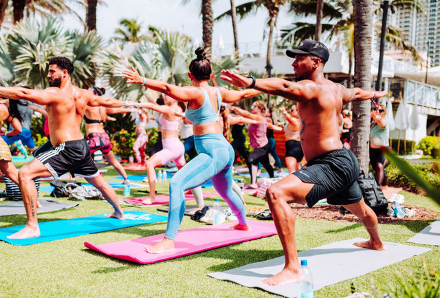 A group of people practicing yoga on mats in a grassy area with palm trees.