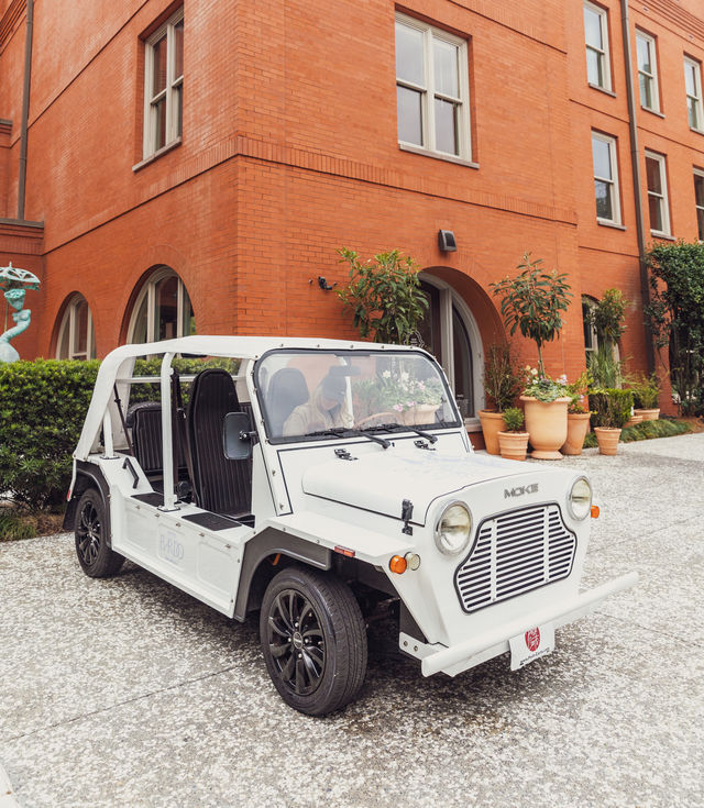 A white car is parked in front of a red brick building with potted plants.