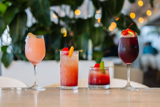 Four colorful cocktails on a table with a blurred background of plants and lights.