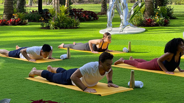 People practice yoga on mats in a grassy area with a statue and tropical plants.