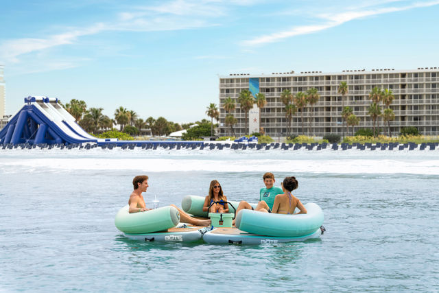 A group of people are sitting on inflatable rafts in the water, with a large hotel building and a water slide in the background.