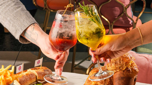 two hands holding wine glasses with drinks and a table with food