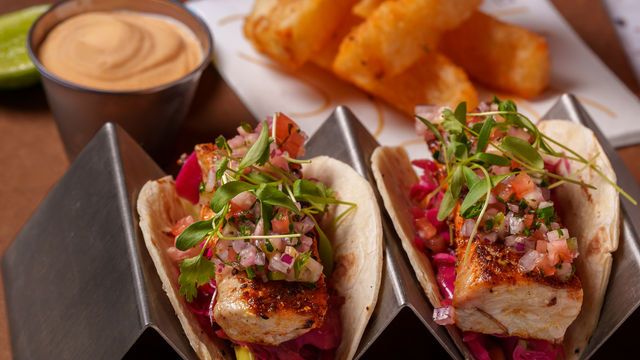 Two tacos with fish, salsa, and microgreens on a metal plate.